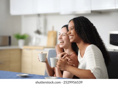 Funny interracial lesbian couple laughing hugging and drinking coffee in the kitchen at home - Powered by Shutterstock