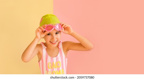 Funny joy girl in striped swimsuit and a Swimming Cap, puts on Swimming Goggles, isolated on an pink and yellow studio background. Learning to swim in the pool from an early age.  Happy baby - Powered by Shutterstock