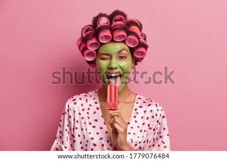 Funny joyful housewife licks delicious ice cream, winks eye, gets pleasure from eating cold refreshing summer dessert, wears green mask on face, hair rollers, dressed in casual outfit, has fun at home