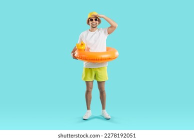 Funny joyful young man set up for summer vacation and looking for good offer from travel agency. Happy guy in summer clothes and with inflatable circle looking into distance on light blue background.