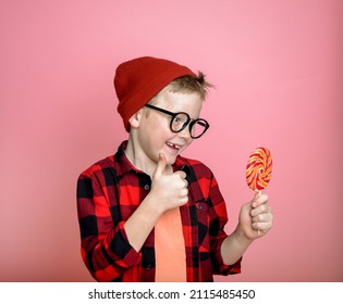 Funny Kid Boy Have Some Fun With Candy Isolated On Pink. Child In Red With Lollipop Shows Thumb Up And Smile. Stylish School Kid Eating Sugar Food