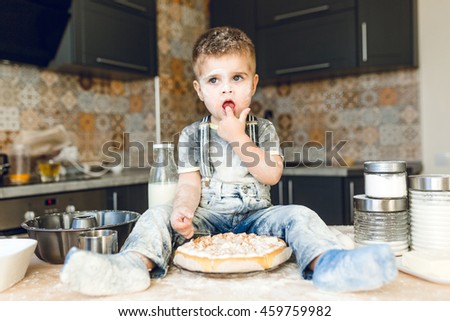 Funny kid sitting on the kitchen table in a roustic kitchen playing with flour and tasting a cake. He is covered in flour and looks funny. He puts his finger in the mouth.