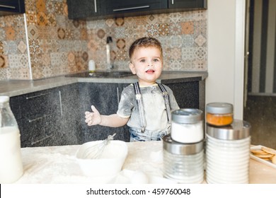 Funny Kid Standing In A Rustic Kitchen Playing With Flour And Throwing It In The Air. He Is Covered In Flour And Looks Funny. He Is Cute. Milk And Different Jars Stand On The Table.