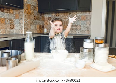 Funny Kid Standing In A Rustic Kitchen Playing With Flour And Throwing It In The Air. He Is Covered In Flour And Looks Funny. He Is Cute. Milk And Different Jars Stand On The Table.