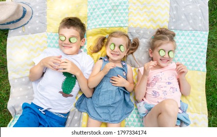 Funny Kids Having Fun During Summer Holidays At Family Camp. Children Try On A Cucumber Mask For Fun Lying On A Quilt. The Concept Of Carelessness And Unschooling. Happy Childhood.