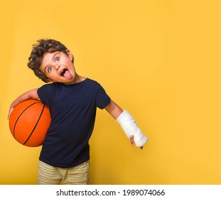 Funny Kittle Boy With Broken Arm With Basketball Isolated On Yellow. Kid Injured After Accident During Summer Vacation In Extreme Park. Child With Wears Cast Ready To Be Healthy And Happy. Sport Time.