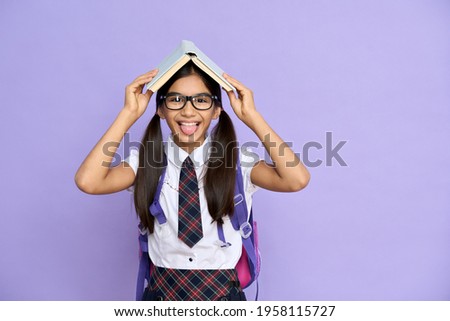 Funny latin schoolgirl in glasses holding book on her head having fun teasing with tongue. Portait of adorable happy indian teenage looking at camera isolated on violet background.