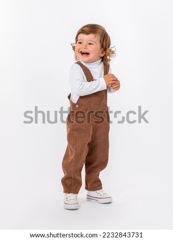 A funny laughing 2-year-old toddler boy with a wide smile with curly hair in a corduroy jumpsuit on a white background in a white jacket and sneakers stands half-side with bent arms.
