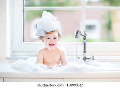 Funny Laughing Baby Girl Splashing Water Playing In A Kitchen Sink Full With Foam Next To A Window