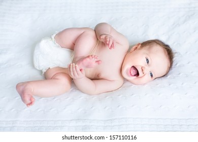Funny laughing baby wearing a diaper playing with her feet on a white knitted blanket