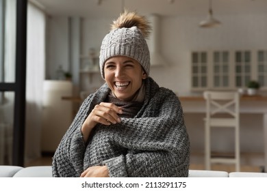 Funny laughing Hispanic woman wear knitted grey bobble hat wrapped in warm plaid sit on sofa relaxing warmup while watch comedy TV show, spend carefree weekend alone at home having fun feel overjoyed