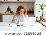 Funny laughing redhead female tutor having video chat with her student, sitting at kitchen table in front of laptop with copy book, coffee cup and smartphone on desk, explaining new topic