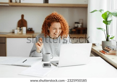 Funny laughing redhead female tutor having video chat with her student, sitting at kitchen table in front of laptop with copy book, coffee cup and smartphone on desk, explaining new topic