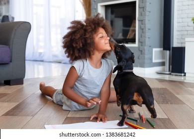 Funny little African American girl lying on floor coloring picture having fun with dog, family pet kissing playing with small child painting at home, kid laugh entertaining with domestic animal
