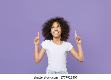 Funny Little African American Kid Girl 12-13 Years Old In White T-shirt Isolated On Violet Wall Background Studio Portrait. Childhood Lifestyle Concept. Mock Up Copy Space. Pointing Index Fingers Up