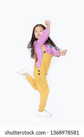 Funny Little Asian Child Girl In Pink-yellow Dungarees Jumping Over White Background. Freedom Kid Movement Concept.