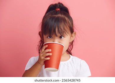 Funny little Asian girl holds disposable paper cup in her hands and drinks water standing on a pink isolated background.  - Shutterstock ID 2365428745