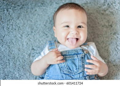 Funny Little Baby Boy In Denim Lying On Grey Blanket. 6 Month Old Child Smiles With His Tongue Out.