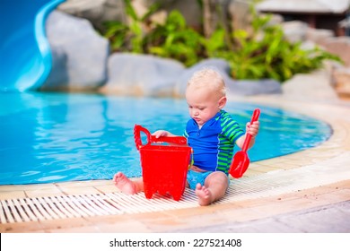 Funny Little Baby Boy In Sun Protection Shirt And Swimming Diaper Playing With A Red Toy Bucket At A Pool In A Tropical Resort