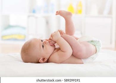 Funny little baby boy wearing a nappy playing with his legs in nursery. Cute kid sucking his feet. Child after bath or shower on bed. Infant skin care.