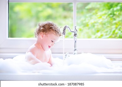 Funny Little Baby Girl With Wet Curly Hair Taking A Bath In A Kitchen Sink With Lots Of Foam Playing With Water Drops And Splashes Next To A Big Window With Garden View