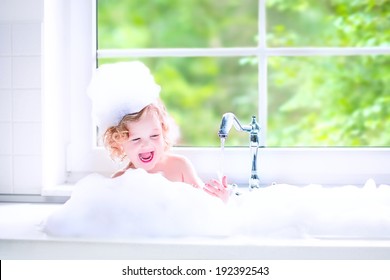 Funny Little Baby Girl With Wet Curly Hair Taking A Bath In A Kitchen Sink With Lots Of Foam Playing With Water Drops And Splashes Next To A Big Window With Garden View