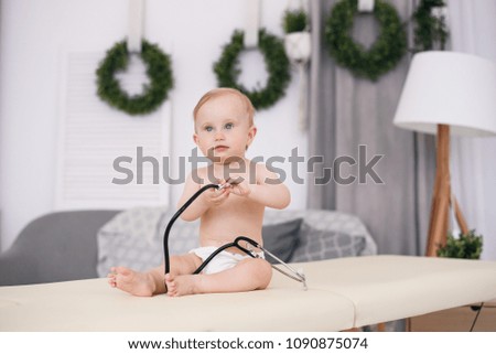 Funny little baby sitting on massage table and playing with stethoscope in modern medical office.