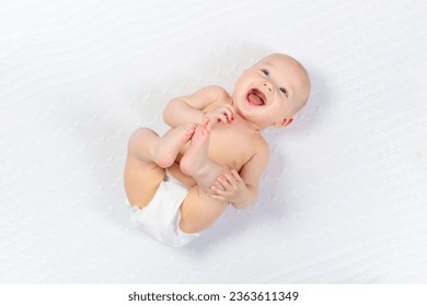 Funny little baby wearing a diaper playing on a white knitted blanket in a sunny nursery. Child after bath or shower on a fresh towel. Infant nappy change and skin care. Cute kid playing with his feet - Powered by Shutterstock