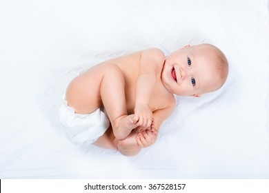 Funny Little Baby Wearing A Diaper Playing On A White Knitted Blanket In A Sunny Nursery. Child After Bath Or Shower On A Fresh Towel. Infant Nappy Change And Skin Care. Cute Kid Playing With His Feet