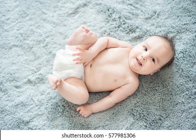 Funny Little Baby Wearing A Diaper Playing On A Grey Blanket In A Sunny Nursery. Child After Bath Or Shower On A Fresh Towel. Infant Nappy Change And Skin Care