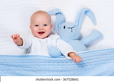 Funny Little Baby Wearing A Warm Knitted Jacket Playing With Toy Bunny Relaxing On White Cable Knit Blanket In Sunny Nursery. Kids Winter Clothing And Bedding. Hand Made Toys And Textile For Children.