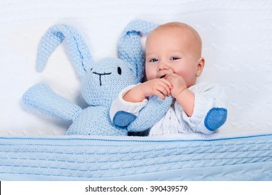 Funny Little Baby Wearing A Warm Knitted Jacket Playing With Toy Bunny Relaxing On White Cable Knit Blanket In Sunny Nursery. Kids Winter Clothing And Bedding. Hand Made Toys And Textile For Children.