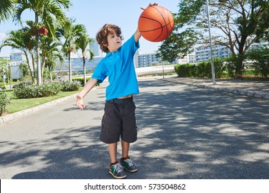 Funny little basketball player with dark curly hair bouncing ball in sunny park with palm trees, full-length portrait