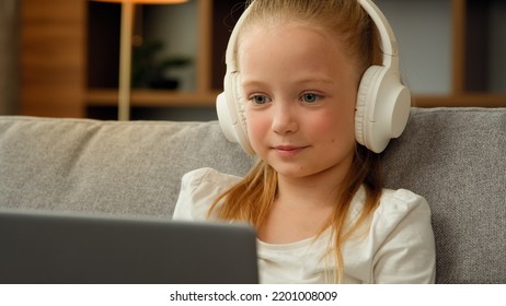 Funny Little Blonde Girl Child Cute Schoolgirl Pretty Beautiful Kid Young Blogger Wears Headphones Sit On Sofa Talking To Webcam Making Online Video Call Recording Vlog Having Fun Waving Hand Greeting