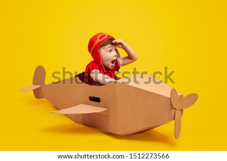 Funny little boy in aviator hat keeping hat near forehead and looking away with amazed face expression while piloting cardboard plane against yellow background