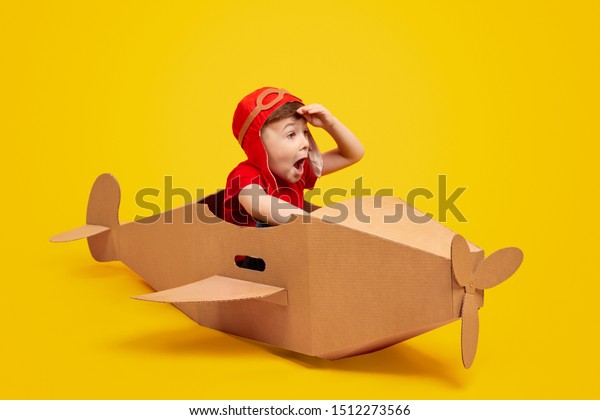 Funny little boy in aviator hat\
keeping hat near forehead and looking away with amazed face\
expression while piloting cardboard plane against yellow\
background