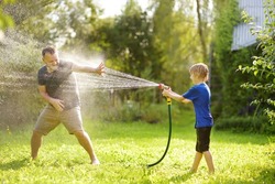 Funny Little Boy With His Father Playing With Garden Hose In Sunny Backyard. Preschooler Child Having Fun With Spray Of Water. Summer Outdoors Activity For Family With Kids.