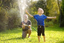 Funny Little Boy With His Father Playing With Garden Hose In Sunny Backyard. Preschooler Child Having Fun With Spray Of Water. Summer Outdoors Activity For Family With Kids. Dad Involved In Parenting