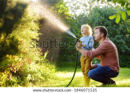 Funny little boy with his father watering plants and playing with garden hose in sunny backyard. Preschooler child having fun with spray of water. Summer outdoors activity for kids.