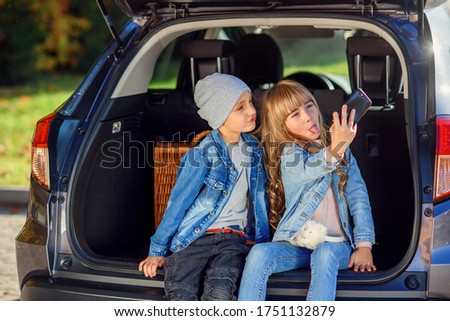 Funny little boy and his pretty fashionable sister with blond hair taking photos on modern smart phone sitting in the opened car's trunk. Family vacation trip by car.