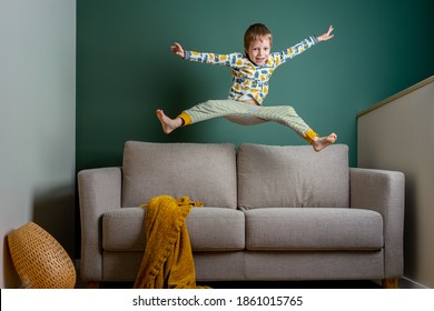 Funny Little Boy Kid Jumping On A Couch And Making A Mess In Modern Interior With Dark Green Wall And Grey Sofa. Copy Space, Horizontal