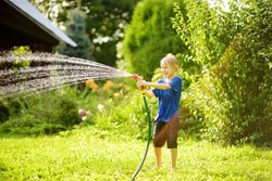 Funny Little Boy Playing With Garden Hose In Sunny Backyard. Preschooler Child Having Fun With Spray Of Water. Summer Outdoors Activity For Family With Kids.