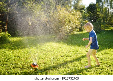 Funny Little Boy Playing With Garden Sprinkler In Sunny Backyard. Preschooler Child Laughing, Jumping And Having Fun With Spray Of Water. Summer Outdoors Activity For Kids.