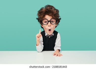 Funny little boy in school uniform and glasses with big head looking at camera with opened mouth, and pointing up while sitting at desk during lesson against turquoise background