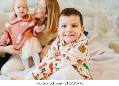 A Funny Little Boy Sits On A Bed With His Mom And Sister. Happy Childhood. Small Children In The Family. Childhood Jealousy And Sibling Relationships.