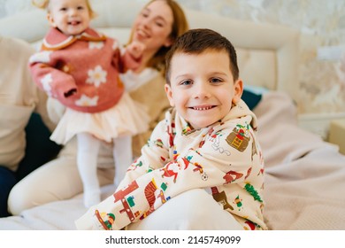 A Funny Little Boy Sits On A Bed With His Mom And Sister. Happy Childhood. Small Children In The Family. Childhood Jealousy And Sibling Relationships.