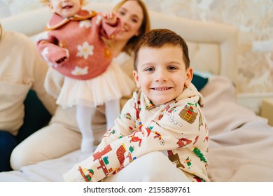 A Funny Little Boy Sits On A Bed With His Mom And Sister. Happy Childhood. Small Children In The Family. Childhood Jealousy And Sibling Relationships.