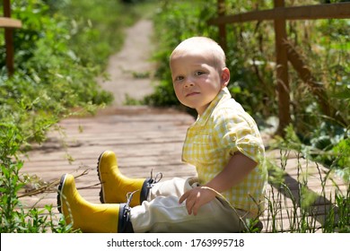 A Funny Little Boy In Yellow Rubber Boots Sits On The Bridge