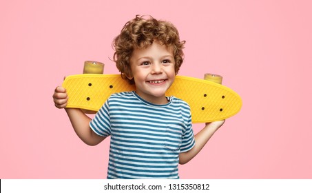 Funny little boy with yellow skateboard smiling and looking away while standing on pink background - Shutterstock ID 1315350812