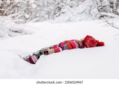 Funny Little Caucasian Girl In A Red Hat And Jumpsuit Is Lying In The Snow In The Winter Forest.Winter Fun,active Lifestyle Concept.Selective Focus,copy Space.